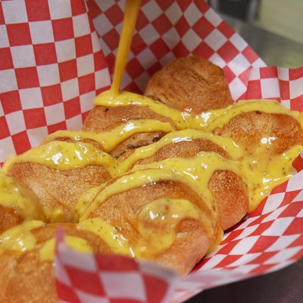 Golden-brown garlic knots are generously drizzled with melted cheese sauce in a red and white checkered basket.