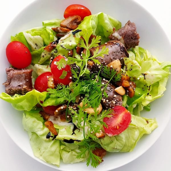Fresh green salad with cherry tomatoes, meat, nuts, and seeds in a white bowl.